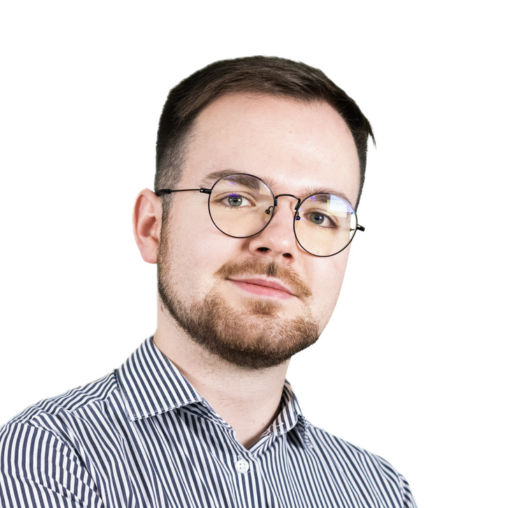 Zdjęcie portretowe. Mężczyzna z brązowymi włosami w okularach i zarostem. Lekko uśmiechnięty.